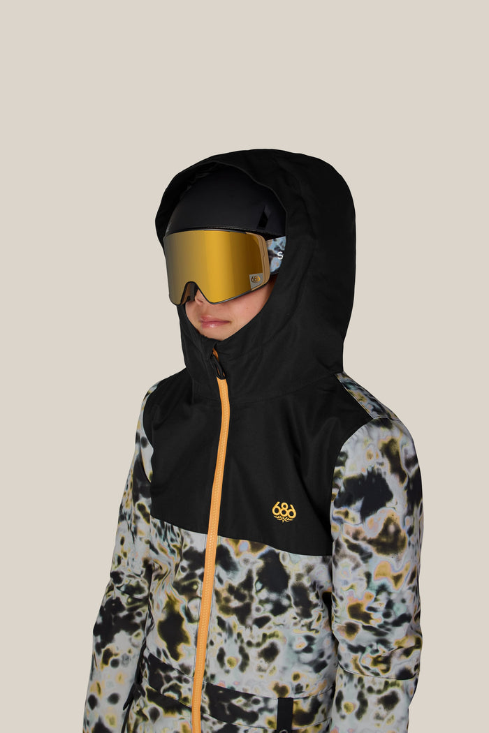 A child wearing the 686 Boysβ Shazam Insulated One-Piece by 686, featuring a black hooded design with a yellow zipper and abstract pattern, paired with large reflective ski goggles and a helmet, stands against a plain beige background.