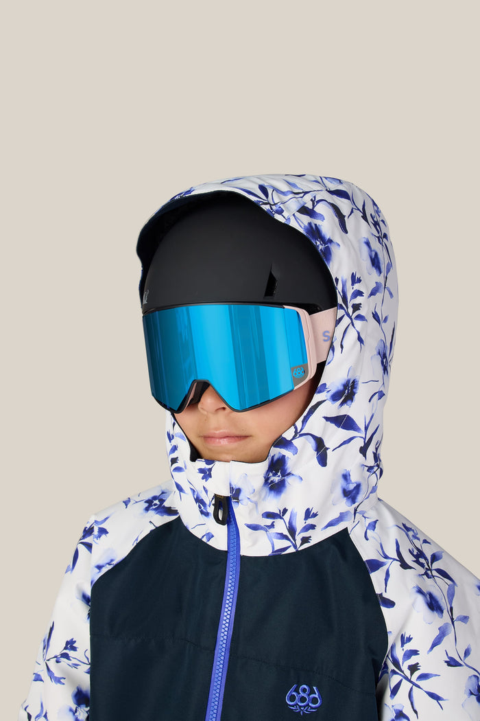 A child wearing the 686 Girlsβ Shine Insulated One-Piece stands against a plain beige background, paired with blue reflective ski goggles and a black helmet.