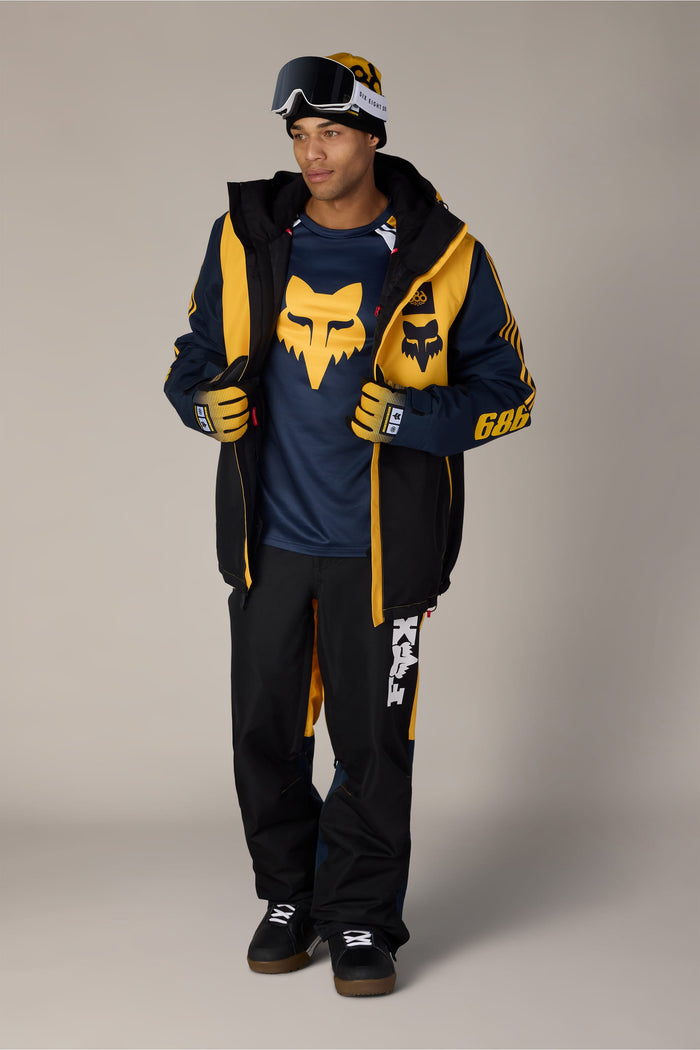 A man wearing a Fox Racing beanie and goggles, a navy and yellow snowboarding jacket, and 686 Menβs Fox Racing Spec Shell Pant.