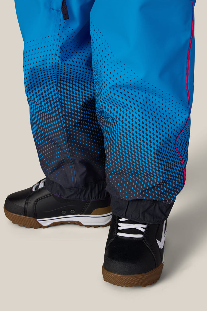 Close-up of a person wearing 686 Womenβs Fox Racing 97 Shell Pants in blue with a black geometric gradient pattern and black snow boots.