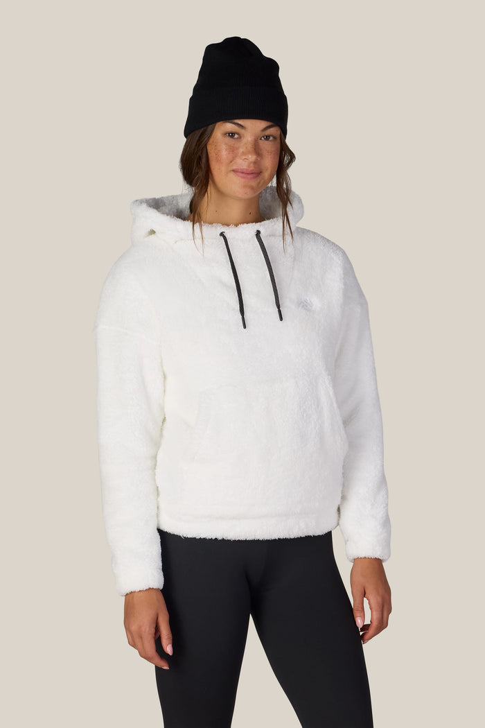 A woman stands facing forward, smiling slightly, against a plain beige background, wearing the 686 Womenβs Sherpa Hoody in white by 686, along with a black beanie and black leggings.