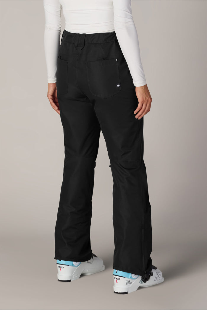 A person is shown from the back, standing on a plain, light-colored background, wearing the 686 Womenβs Slim Line Pant in black polyester waterproof fabric, paired with a white long-sleeve top and white ski boots.