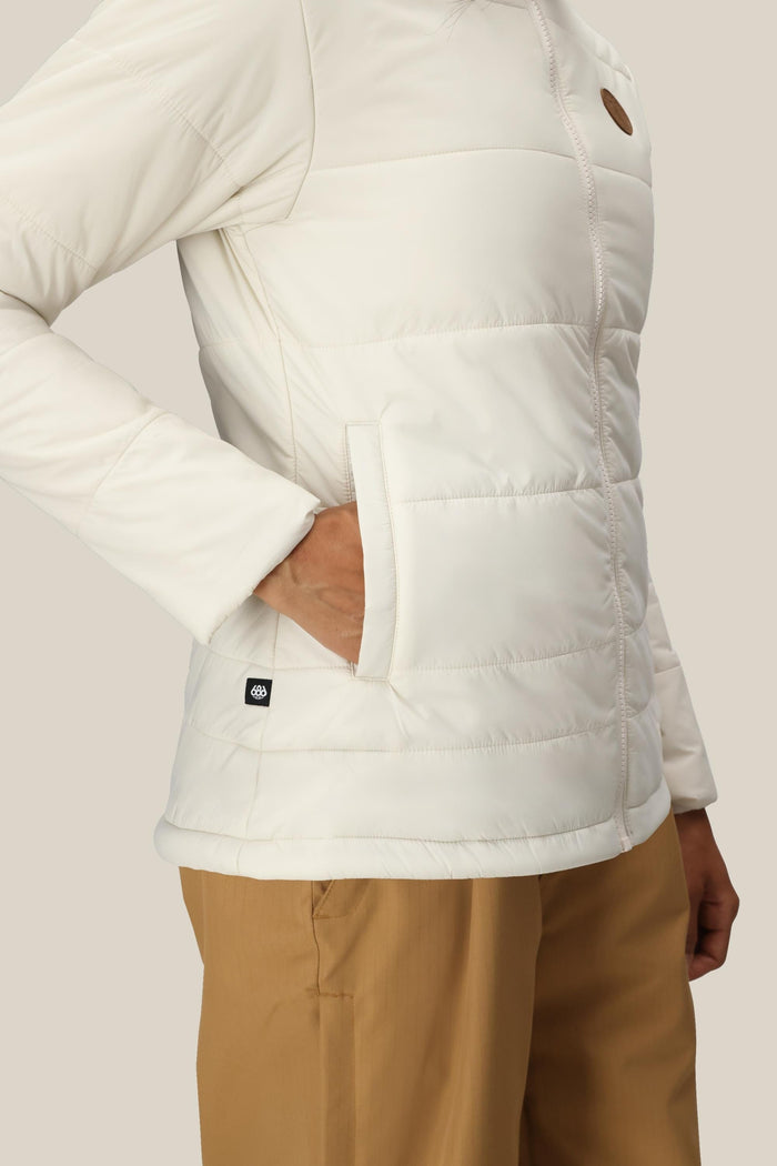A woman wearing the 686 Womenβs Thermal Puff Insulated Jacket with one hand in the pocket and tan pants stands against a neutral background.