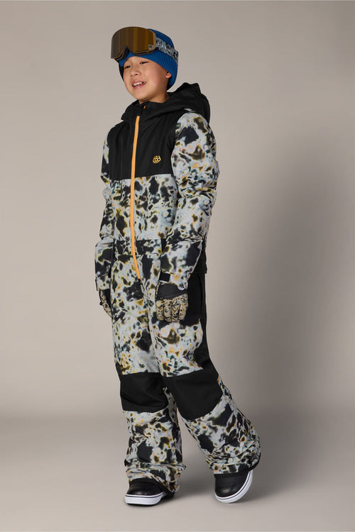 A child smiles against a plain background while wearing the 686 Boys’ Shazam Insulated One-Piece by 686, paired with a blue beanie and ski goggles.