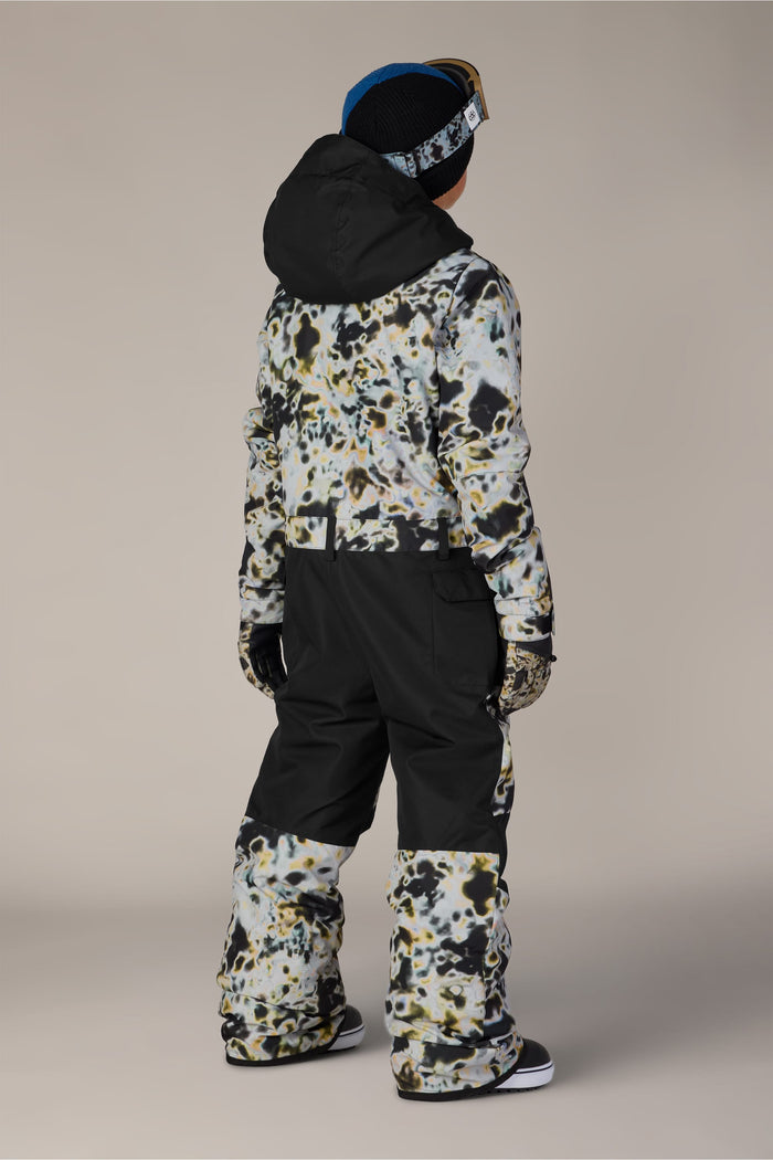 A child faces away, dressed in the 686 Boys’ Shazam Insulated One-Piece by 686, paired with black snow pants, gloves, a black hat, and blue ski goggles against a plain neutral background.