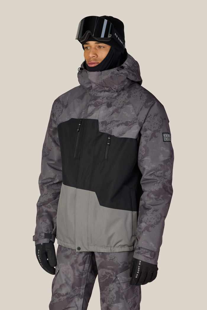 A person wearing the 686 Men’s Geo™ Insulated Jacket in a gray and black camo pattern stands against a plain light background, also dressed in matching pants, black gloves, a black hat, and ski goggles.