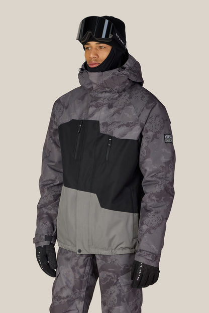A person wearing the 686 Men’s Geo™ Insulated Jacket in a gray and black camo pattern stands against a plain light background, also dressed in matching pants, black gloves, a black hat, and ski goggles.
