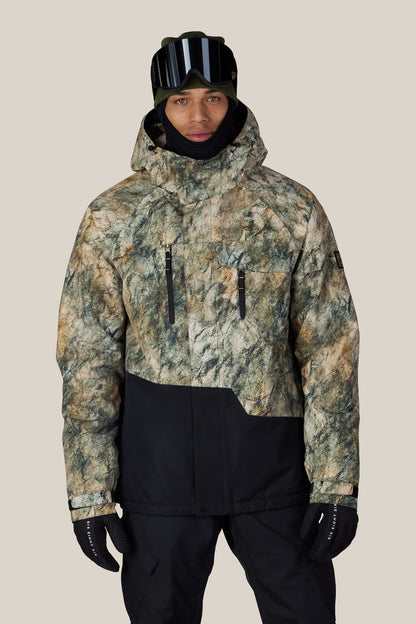 Wearing the 686 Men’s Geo™ Insulated Jacket, a person stands against a plain light background. The jacket features a green and brown camouflage pattern, paired with black snow pants, gloves, ski goggles, and a hood.