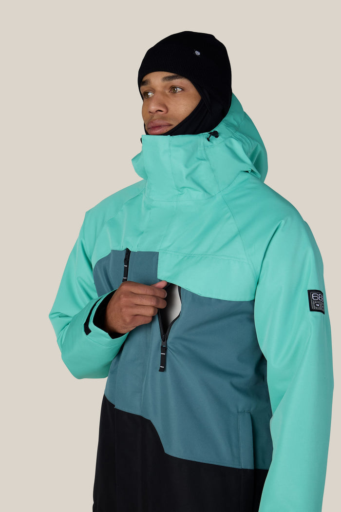 A person wearing the 686 Men’s Geo™ Insulated Jacket in teal and dark green, along with a black beanie, zips up the front while facing slightly to the right against a plain light background.