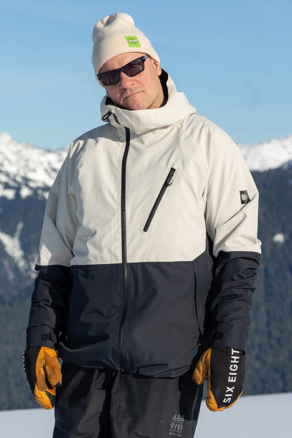 Wearing the 686 Men’s Hydra™ Thermagraph® Jacket, a person stands outdoors against a backdrop of snowy mountains and blue sky, completed with a tan beanie, orange gloves, and sunglasses.