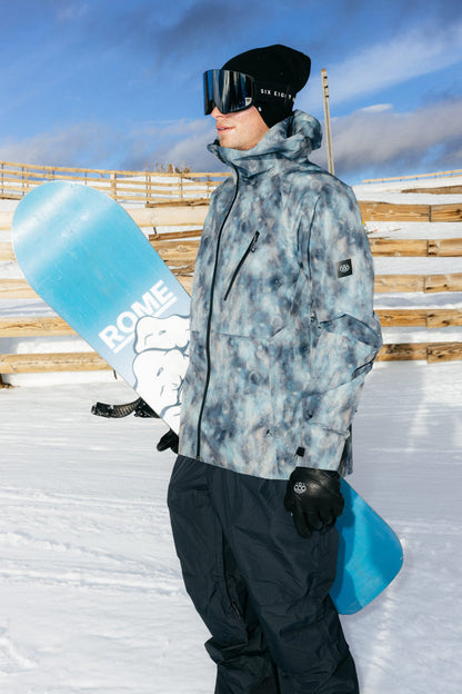 A person wearing the 686 Men’s Hydra™ Thermagraph® Jacket stands on snow with goggles, holding a blue snowboard labeled ROME, against a backdrop of wooden fences and a blue sky.