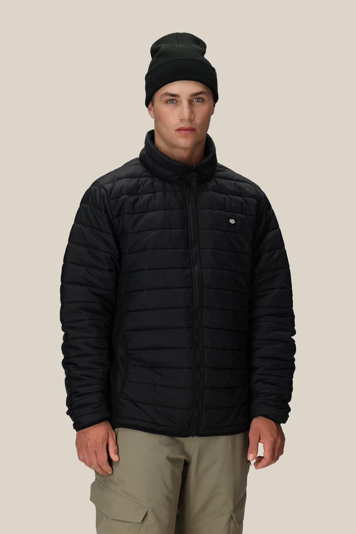 A person wearing the 686 Men’s Thermal Puff Jacket in black, paired with a black beanie and beige pants, stands facing forward against a plain beige background.