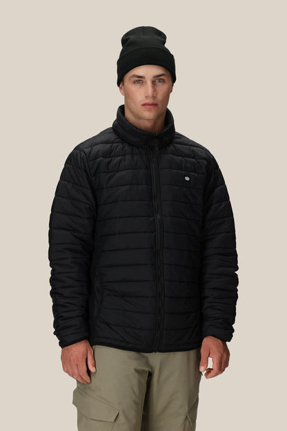 A person wearing the 686 Men’s Thermal Puff Jacket in black, paired with a black beanie and beige pants, stands facing forward against a plain beige background.