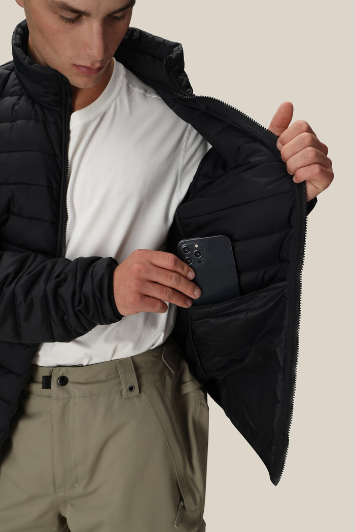 A person in a white shirt and khaki pants puts a black smartphone into the inner pocket of a black 686 Men’s Thermal Puff Jacket by 686, which is unzipped to reveal the pocket.