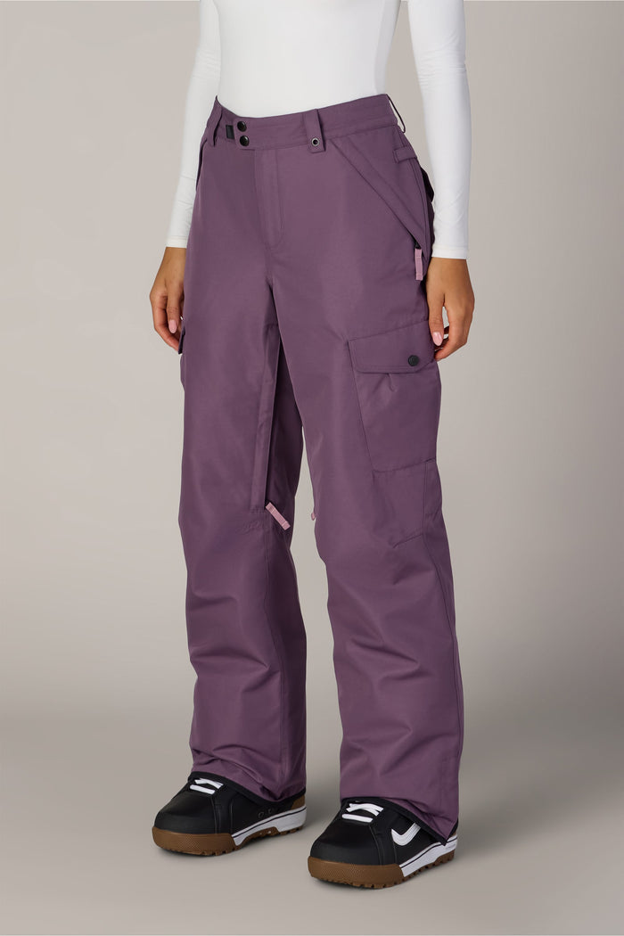 A person wearing the 686 Women’s Aura™ Insulated Cargo Pant in purple, paired with a white long sleeve top and black winter boots featuring white and brown soles, stands against a plain light background.