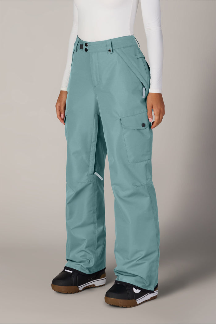 A person wears the 686 Women’s Aura™ Insulated Cargo Pant in light blue, paired with a white long-sleeve top and black snow boots, standing against a plain, light background. Only the lower body is visible.