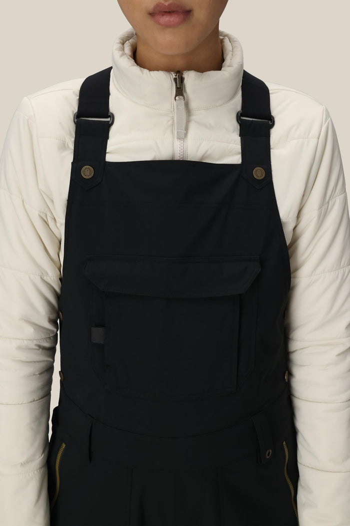 A person is shown from shoulders to waist against a plain background, wearing the 686 Women’s Black Magic™ Insulated Bib over a white zip-up jacket.