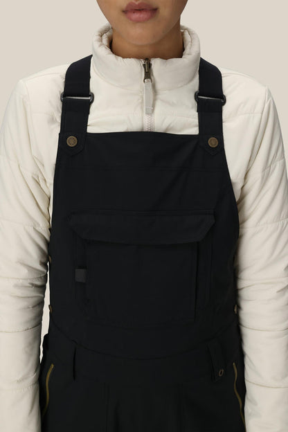 A person is shown from shoulders to waist against a plain background, wearing the 686 Women’s Black Magic™ Insulated Bib over a white zip-up jacket.