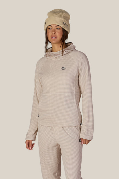 A woman wearing a beige beanie and pants, styled with the 686 Women’s Mission Grid™ Hoody by 686 and a women’s polyester jacket, stands against a plain light background.