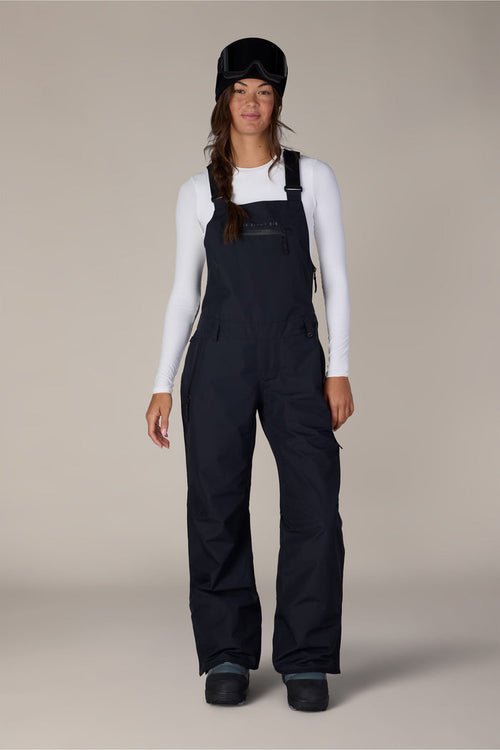 A person wearing a white long-sleeve shirt, the 686 Women’s Regalia™ Bib snow pants in black, black snow boots, and a black ski helmet with goggles stands against a plain background.