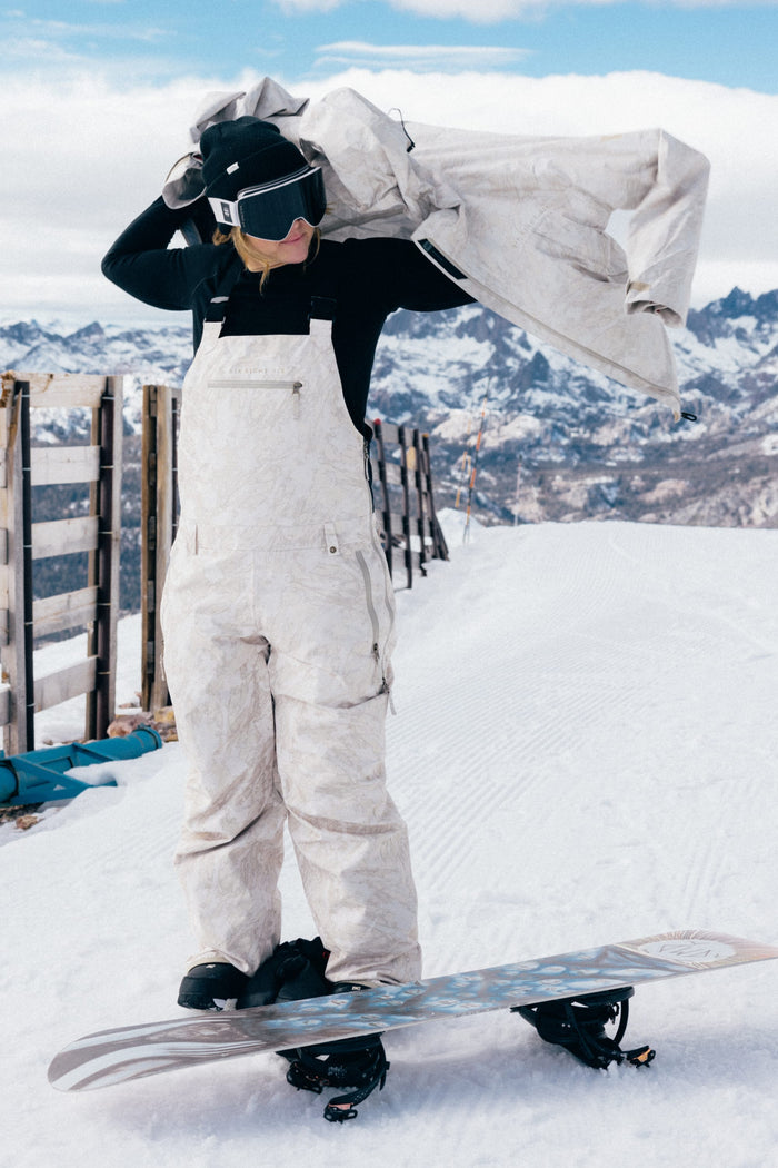 Wearing the 686 Women’s Regalia™ Bib, a person in white snow gear and goggles stands on a snowboard, adjusting their jacket with snowy mountains and a wooden fence in the background.