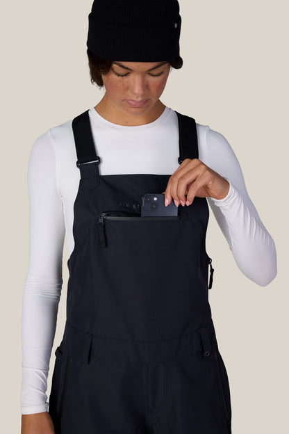A person wearing a white long-sleeve shirt, the 686 Women’s Regalia™ Bib by 686 in black, and a black beanie places a small black device into the zippered chest pocket of the overalls, against a light beige background.