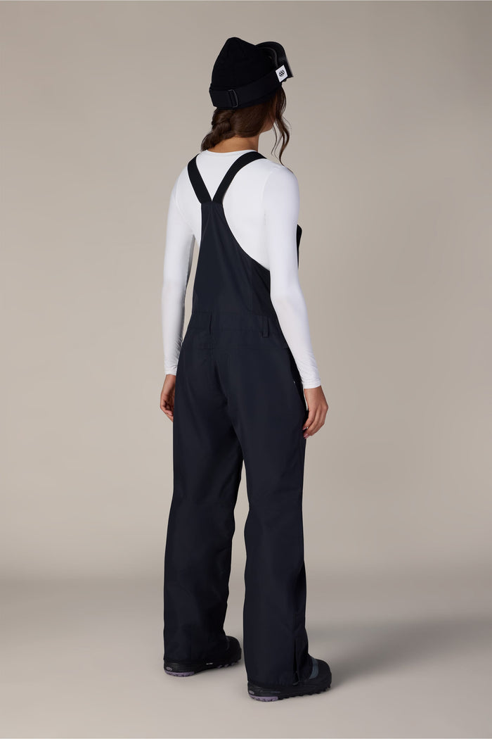 A person stands facing away, wearing a black beanie, a white long-sleeve shirt, and the 686 Women’s Regalia™ Bib by 686—black polyester bib overalls with wide straps. The background is plain and neutral.