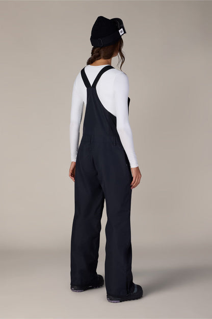 A person stands facing away, wearing a black beanie, a white long-sleeve shirt, and the 686 Women’s Regalia™ Bib by 686—black polyester bib overalls with wide straps. The background is plain and neutral.