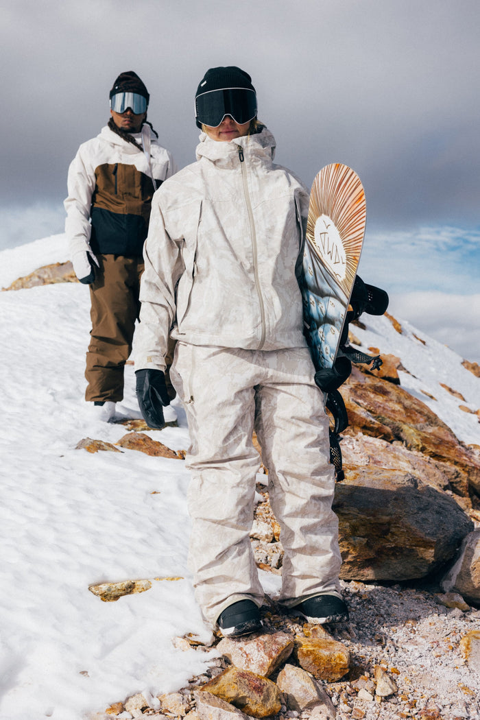 Clad in the 686 Women’s Regalia™ Bib for extra protection against the elements, two people in winter gear stand on a snowy, rocky slope. The person in front holds a snowboard, and both wear goggles, gloves, and hoods. With cloudy skies overhead, they look ready to take on the wintry cold—thanks to their waterproof bibs from 686.