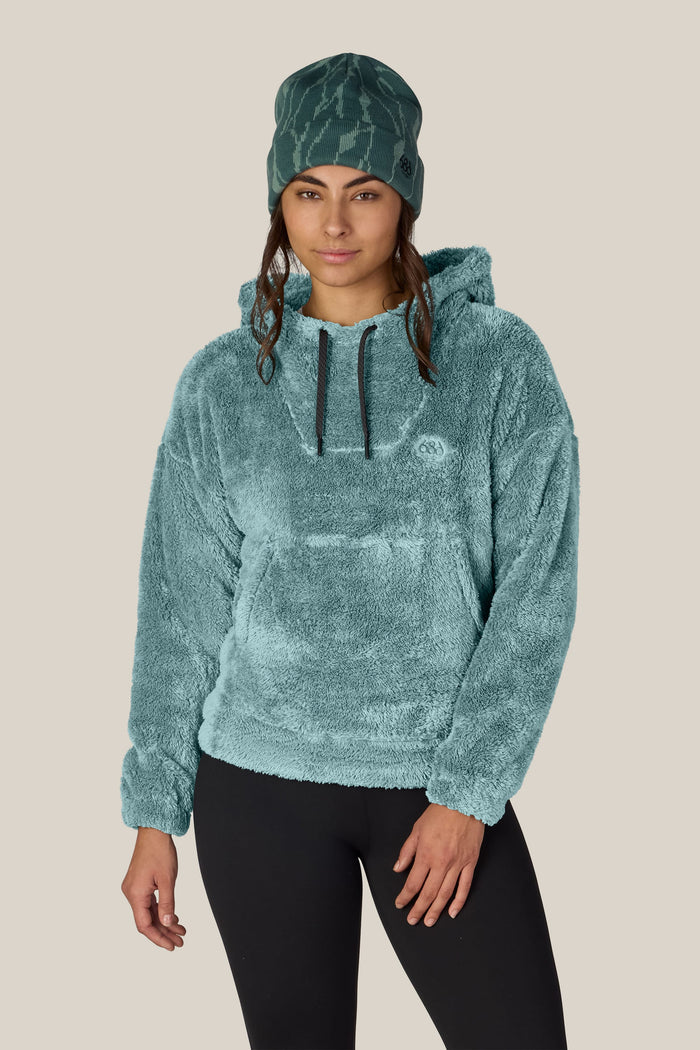 A woman wears the 686 Women’s Sherpa Hoody in teal, paired with black leggings and a matching teal beanie. She stands facing forward against a plain beige background, looking relaxed with one hand by her side.
