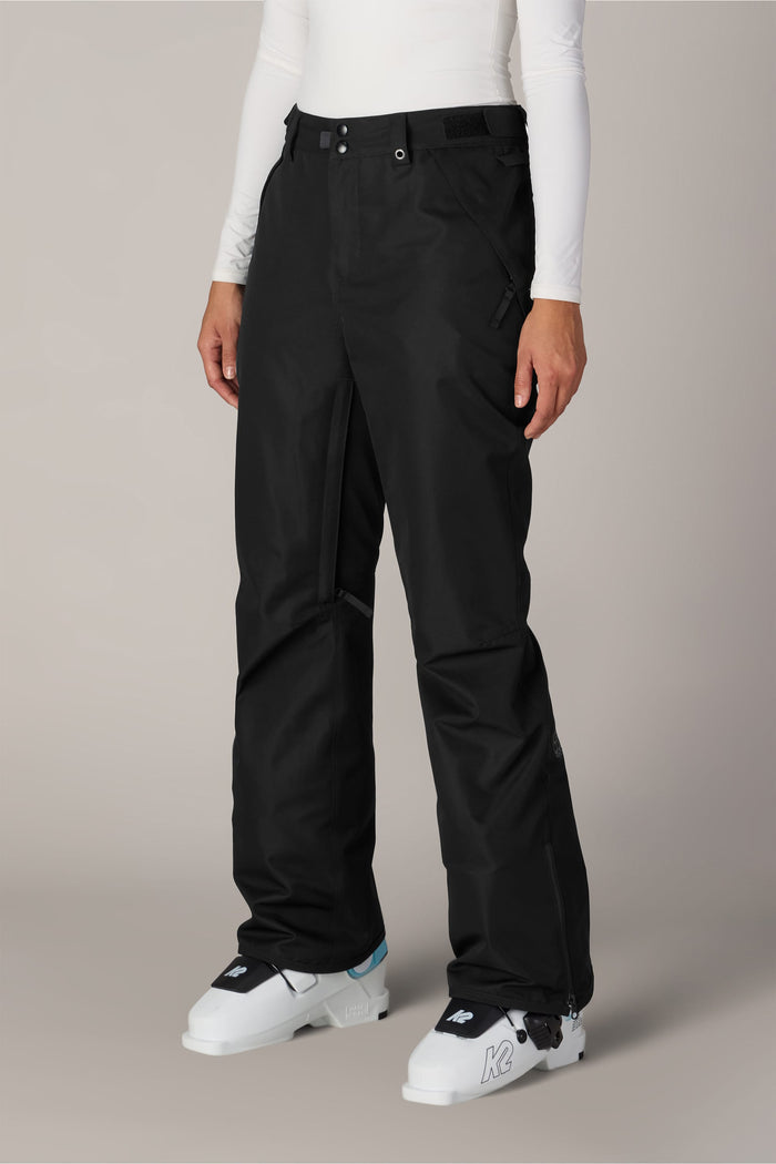 A person is wearing the 686 Women’s Slim Line Pant in black polyester, paired with a white long-sleeve top and white ski boots, standing against a plain light background.