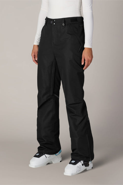A person is wearing the 686 Women’s Slim Line Pant in black polyester, paired with a white long-sleeve top and white ski boots, standing against a plain light background.