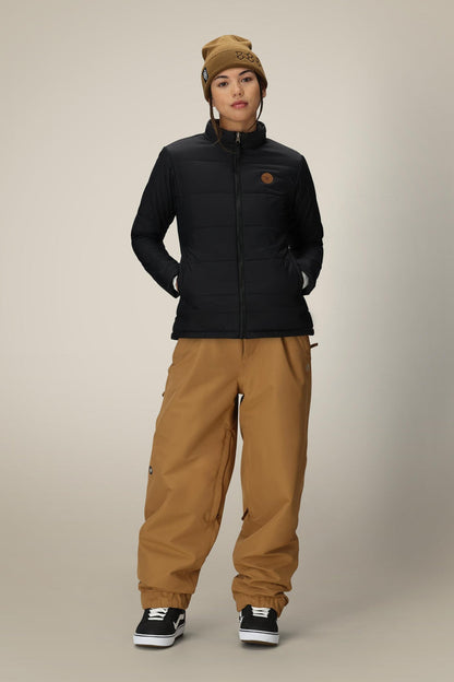 Wearing the 686 Women’s Thermal Puff Insulated Jacket by 686, a person stands with hands in pockets against a plain background, paired with a brown beanie, brown baggy pants, and black sneakers.