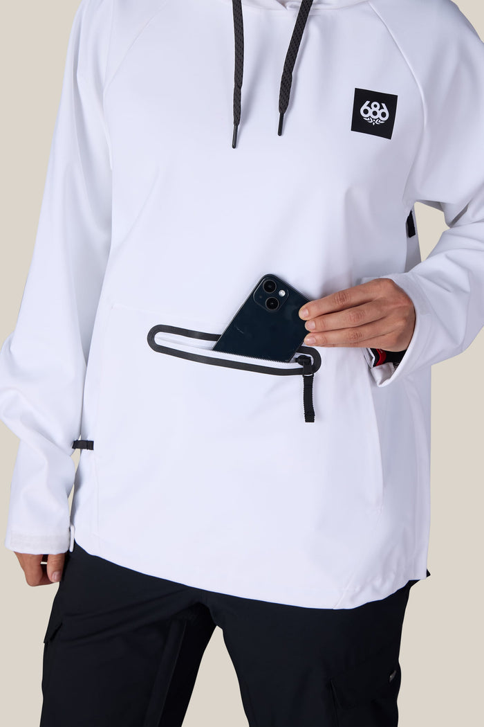 A person wearing the 686 Women’s Waterproof Hoody by 686, featuring black drawstrings and a front zipper pocket, places a black smartphone inside the pocket. Only their torso and one hand are visible.