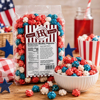 A bag of Poppin' IR American Medley Popcorn stands on a wooden surface with a white bowl, scattered popcorn pieces, a red-and-white striped cup, and patriotic props in the background. The product packaging is clear with a large front label and the brand logo visible.