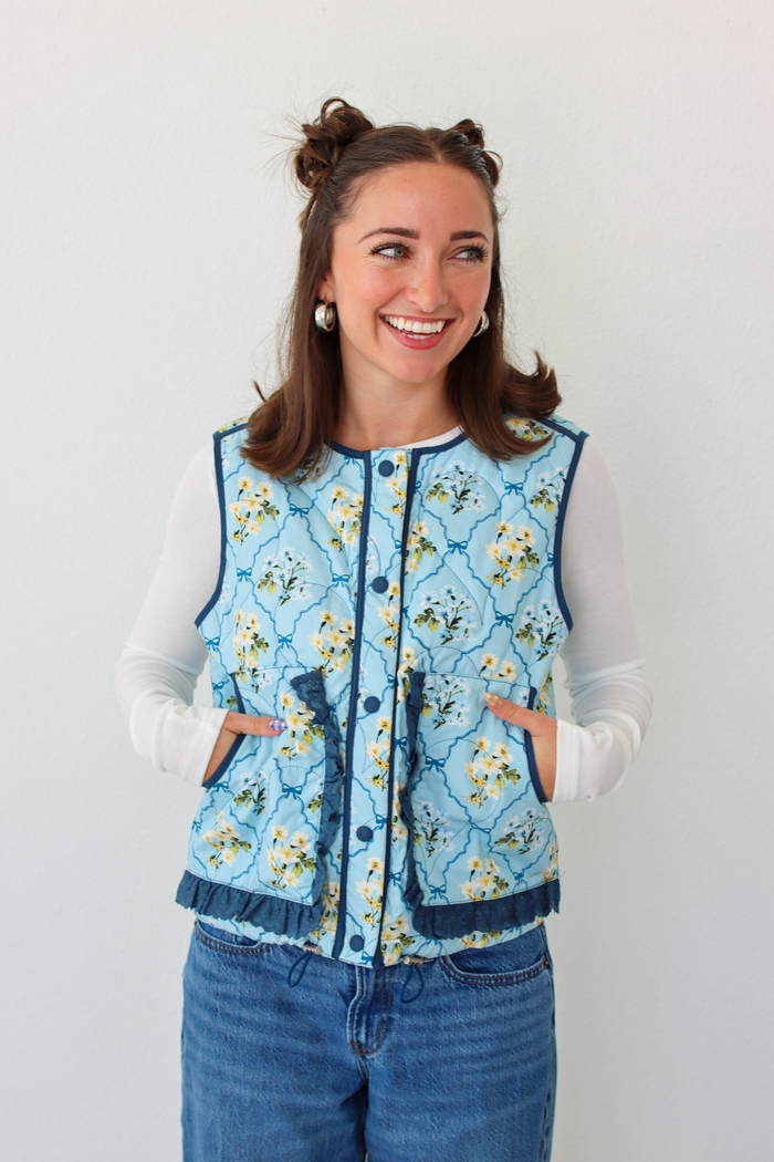 Woman wearing a blue floral puffer vest with a white long sleeve shirt and blue denim jeans