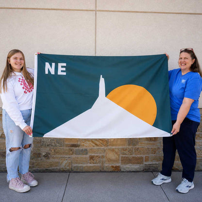 Two women holding a dark green flag with a white mountain, a yellow sun, and the letters 'NE' in white, standing outdoors against a beige wall and stone base.