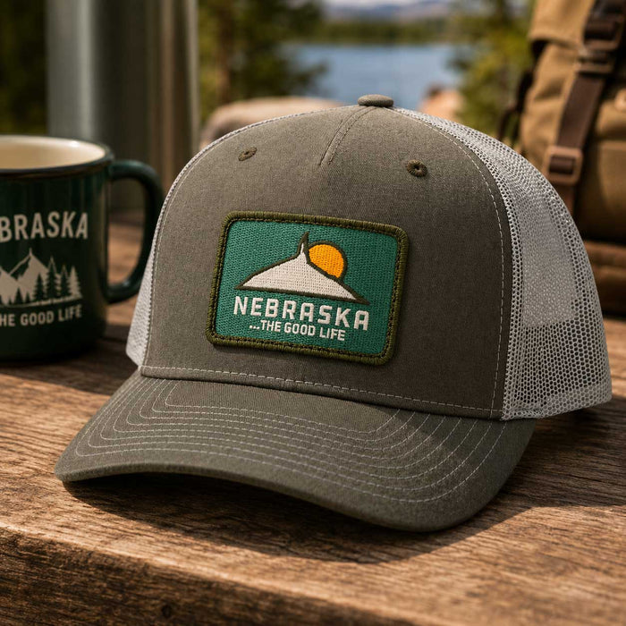 A gray trucker hat with a Nebraska patch on the front, resting on a wooden table next to a green mug with a lake background.