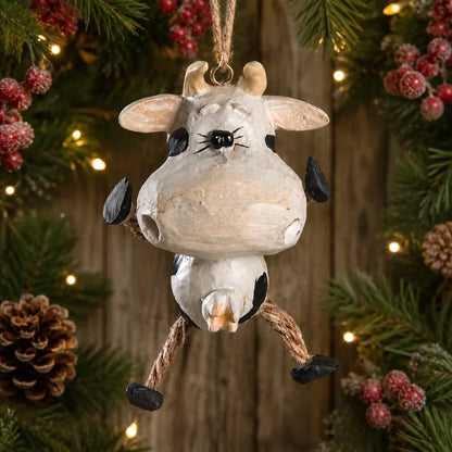 A rustic Christmas hanging ornament shaped like a cow, with a distressed off-white body, black spots, small horns, and rope legs, displayed against a festive holiday background of pine branches, berries, and warm string lights.