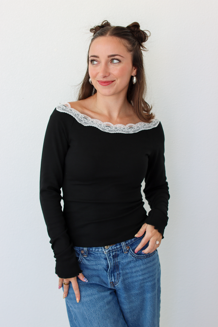 A woman with her hair in two buns is wearing a black off-the-shoulder long-sleeve top with a white lace trim and blue jeans, standing against a plain white wall.