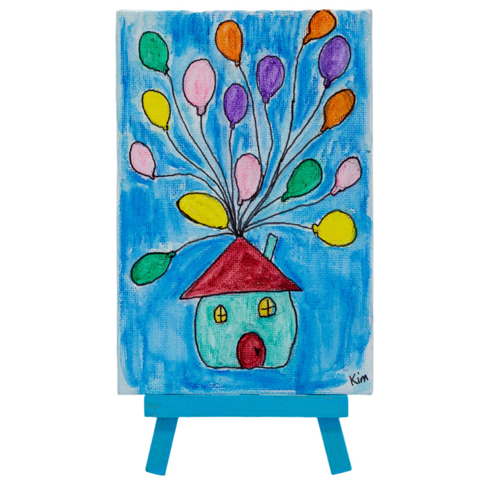A colorful canvas painting featuring a house with a red roof and multiple balloons in various colors released from the top of the house, set against a blue background. The painting is displayed on a small easel.