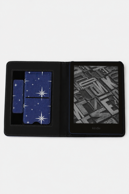 A black leather Kindle e-reader case shown open on a white background, featuring a blue starry night sky pattern on the card slots and a black-and-white book cover displayed on the screen.
